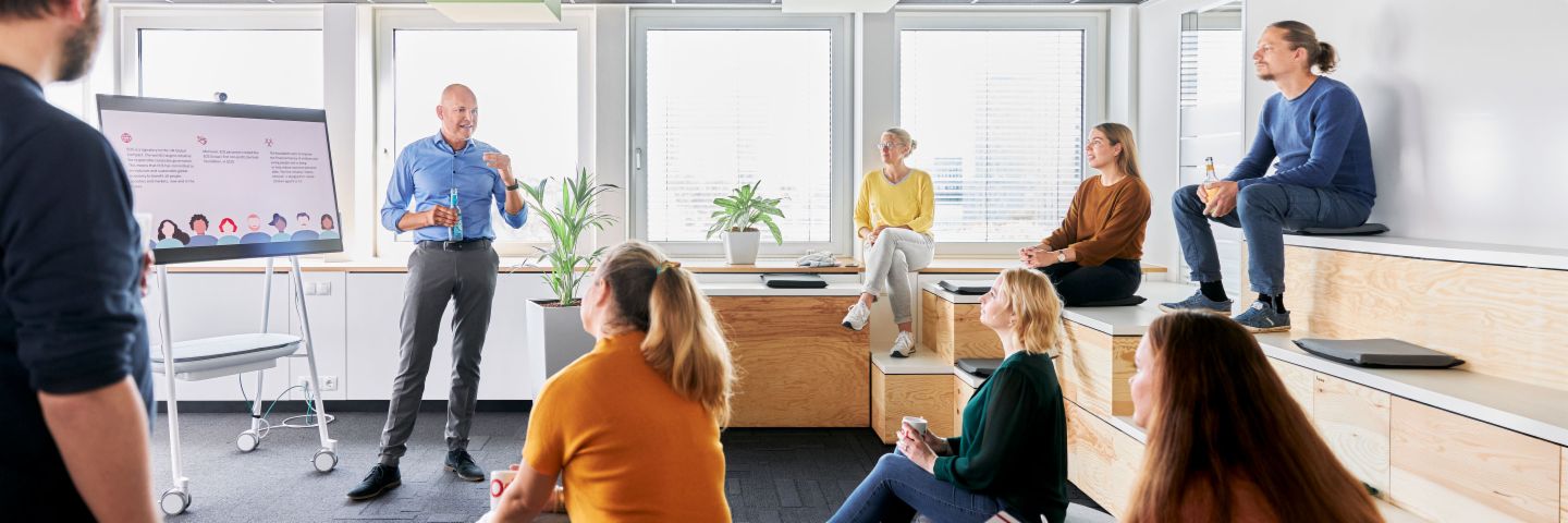 Ein Team in einem modernen Collaborationsbereich eines Büros nimmt an einer Präsentation teil, bei der ein EOS Mitarbeiter vor einem digitalen Whiteboard zu branchenspezifischen Lösungen im Forderungsmanagement spricht.