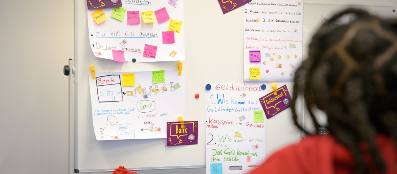 Ein Foto zeigt Post-its und Zettel mit Notizen an einer Tafel, auf denen Begriffe wie „Schulden“, „Bargeld“, „Bank“ und „Geldautomat“ stehen. Darunter steht der Text: „Im Programm ManoMoneta der EOS eigenen finlit foundation können Schüler im Alter von 9 bis 13 Jahren Wissen über Finanzprodukte erwerben.“