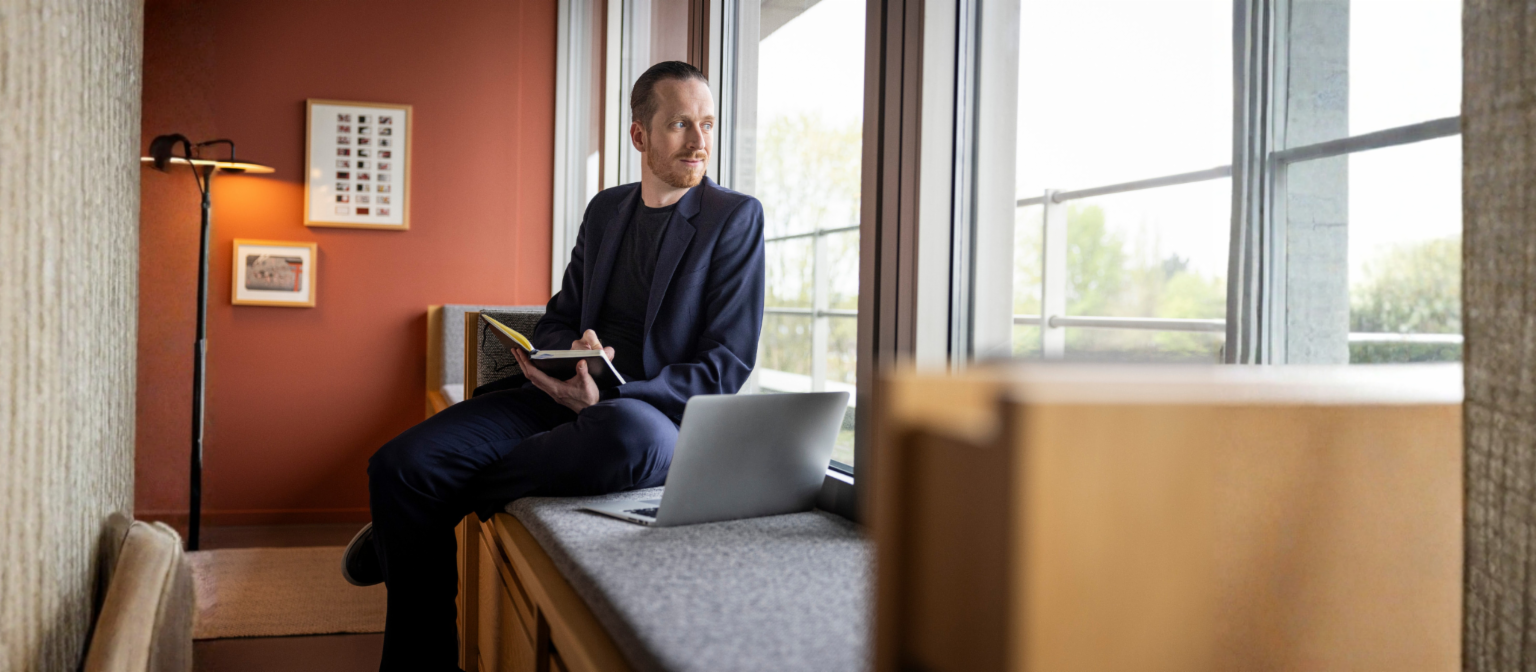 Sebastian Pollmer sitzt mit einem aufgeklappten Laptop und einem Notizbuch in der Hand auf der Fensterbank und schaut aus dem Fenster.