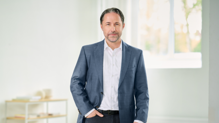 Marwin Ramcke, CEO, steht mit blauem Anzug und der rechten Hand in der Hosentasche in einem hellen Raum. 