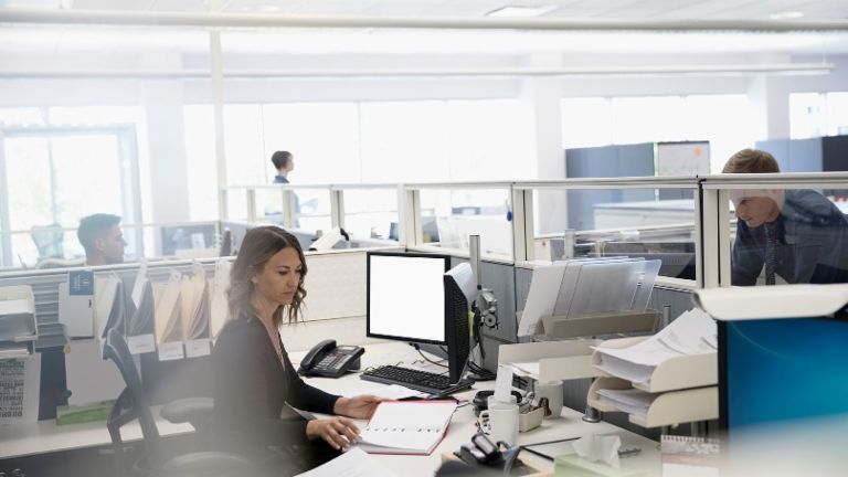 Digitalisierung: Drei Mitarbeiter sind in einem Großraumbüro, eine Frau sitzt zwischen Papieren und ihrem Computer