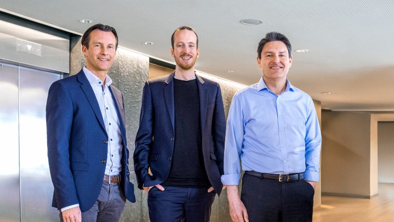 Drei männliche Mitglieder der Geschäftsleitung der EOS Gruppe stehen in Businesskleidung vor Fahrstühlen in modernem Ambiente. 