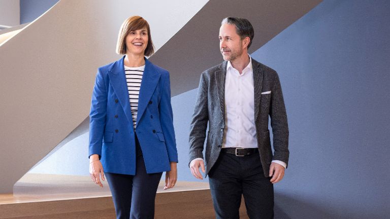Der CEO im grauen Blazer und die CFO der EOS Gruppe in blauem Blazer gehen in einem modernen Büro. Der Raum hat große Fenster, eine geschwungene Wand und stilvolle hängende Lampen.