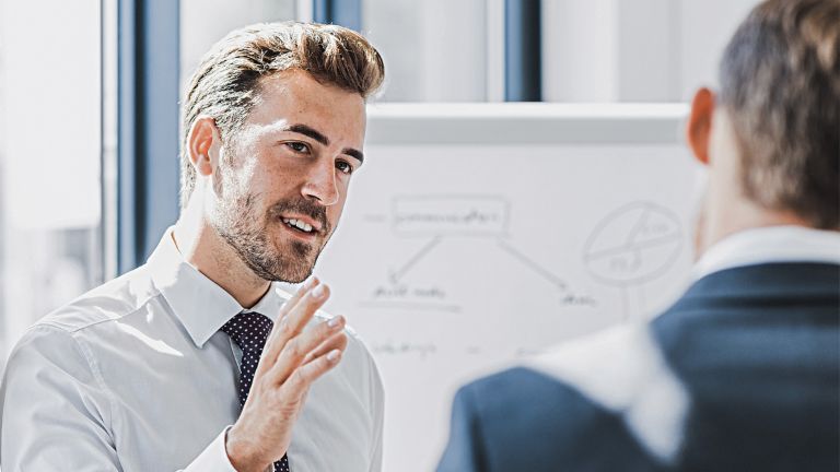Zu sehen ist ein Mann in Business-Bekleidung, der mit freundlich-engagiertem Ausdruck etwas erklärt. Vor ihm steht eine andere Person, die offensichtlich Zuhörer ist. Im Hintergrund ist ein White-Board mit einer Skizze zu sehen.