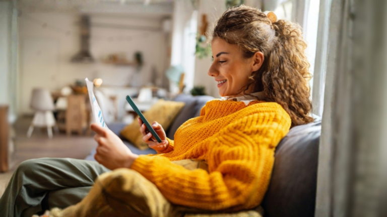 Eine lächelnde junge Frau mit lockigen Haaren in einem gemütlichen gelben Pullover sitzt auf dem Sofa in ihrem Wohnzimmer und schaut auf ihr Smartphone, während sie ein Papierdokument in der Hand hält.