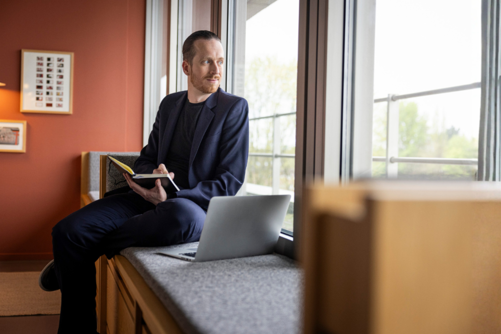 Sebastian Pollmer sitzt mit einem aufgeklappten Laptop und einem Notizbuch in der Hand auf der Fensterbank und schaut aus dem Fenster.