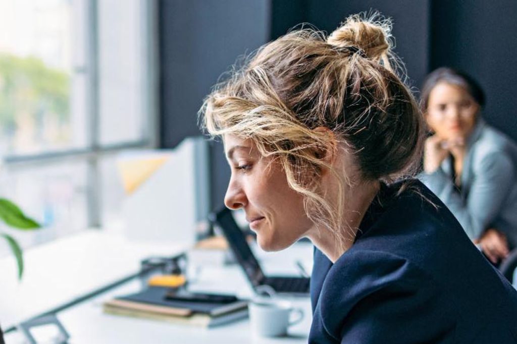 Das Bild zeigt eine Frau, die an einem Schreibtisch in einem modernen Büro sitzt, einen dunklen Blazer trägt und sich auf ihren Laptop konzentriert, während eine Pflanze und einige Büromaterialien auf dem Tisch sichtbar sind und im Hintergrund eine andere Person sitzt.