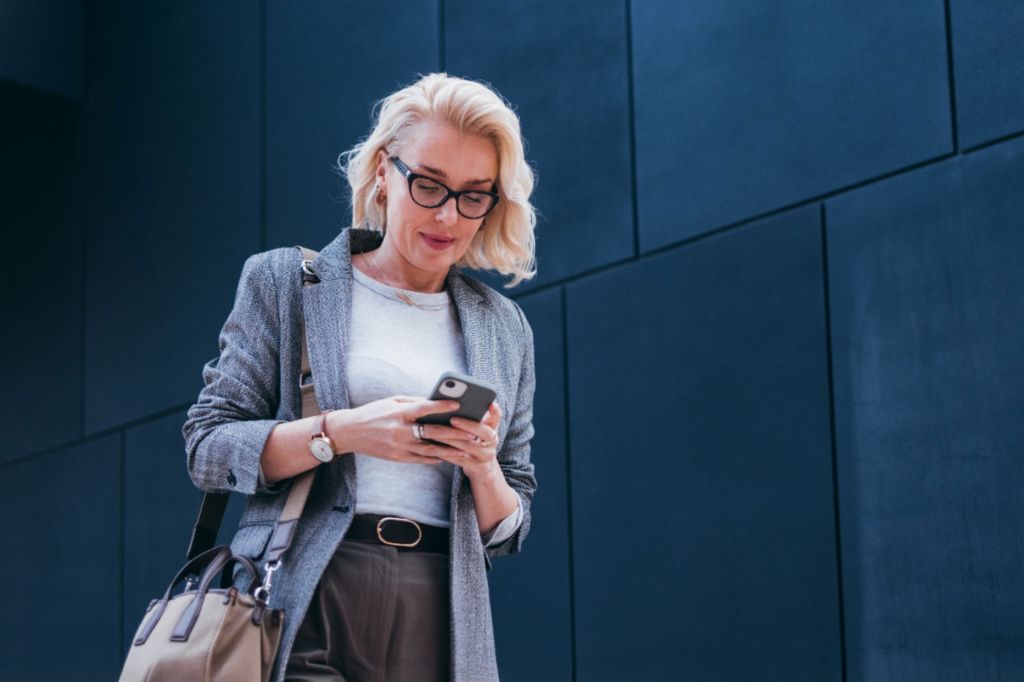 Eine blonde Geschäftsfrau im grauen Blazer steht vor einem modernen Gebäude und schaut lächelnd auf ihr Smartphone.