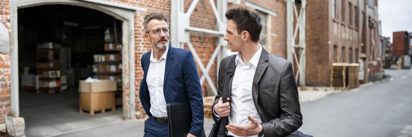 Two businessmen walk side by side past a brick building and have an animated conversation.