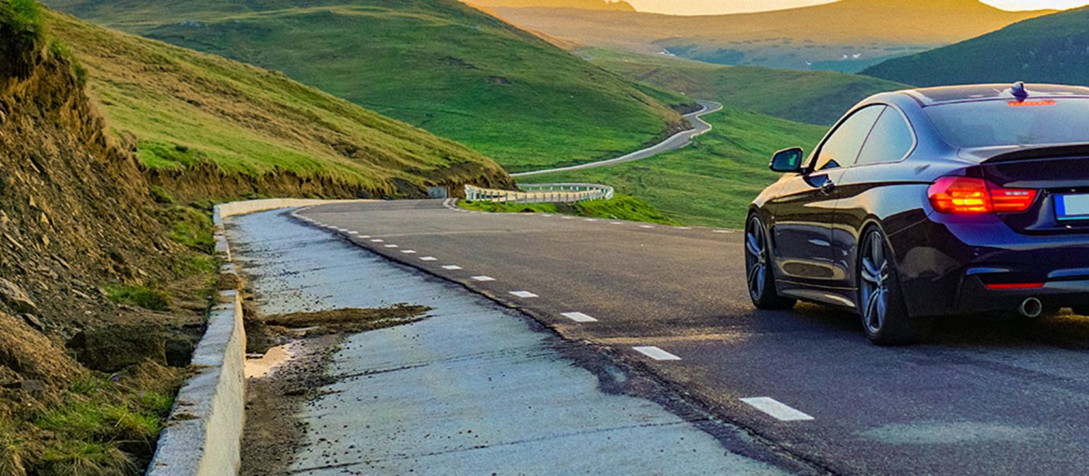 Eine Fotomontage zeigt ein Auto, das auf der rechten Spur einer asphaltierten Straße durch eine grüne, weite Landschaft in Richtung Sonnenuntergang fährt. Auf der entgegenkommenden Spur ist der Asphalt vollständig abgetragen.