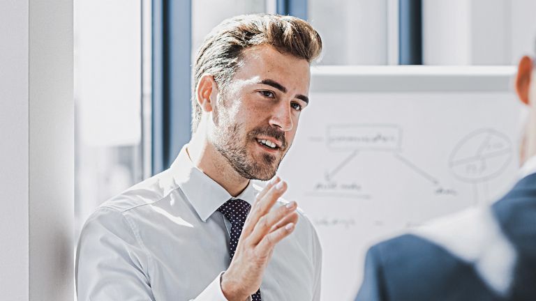 Zu sehen ist ein Mann in Business-Bekleidung, der mit freundlich-engagiertem Ausdruck etwas erklärt. Vor ihm steht eine andere Person, die offensichtlich Zuhörer ist. Im Hintergrund ist ein White-Board mit einer Skizze zu sehen.