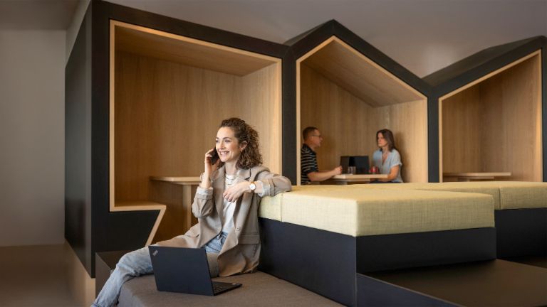 Lächelnde Frau sitzt in einer modernen Büro-Lounge, telefoniert und arbeitet am Laptop. Im Hintergrund arbeiten zwei weitere Personen.