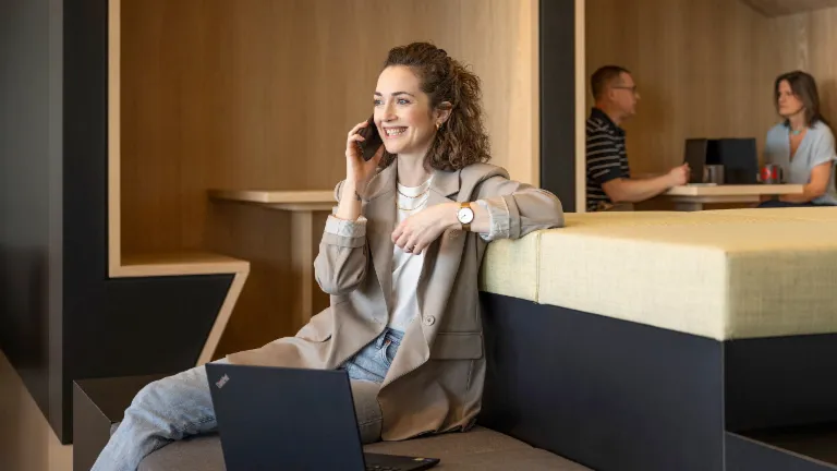 Lächelnde Frau sitzt in einer modernen Büro-Lounge, telefoniert und arbeitet am Laptop. Im Hintergrund arbeiten zwei weitere Personen.
