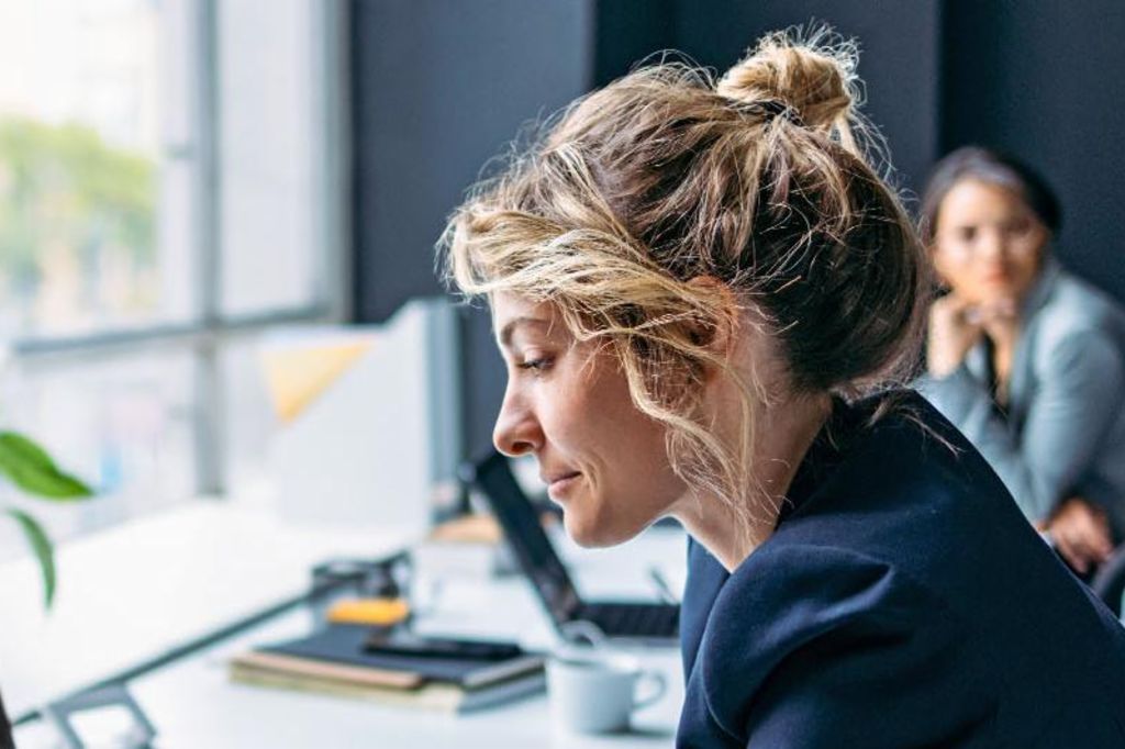 Das Bild zeigt eine Frau, die an einem Schreibtisch in einem modernen Büro sitzt, einen dunklen Blazer trägt und sich auf ihren Laptop konzentriert, während eine Pflanze und einige Büromaterialien auf dem Tisch sichtbar sind und im Hintergrund eine andere Person sitzt.