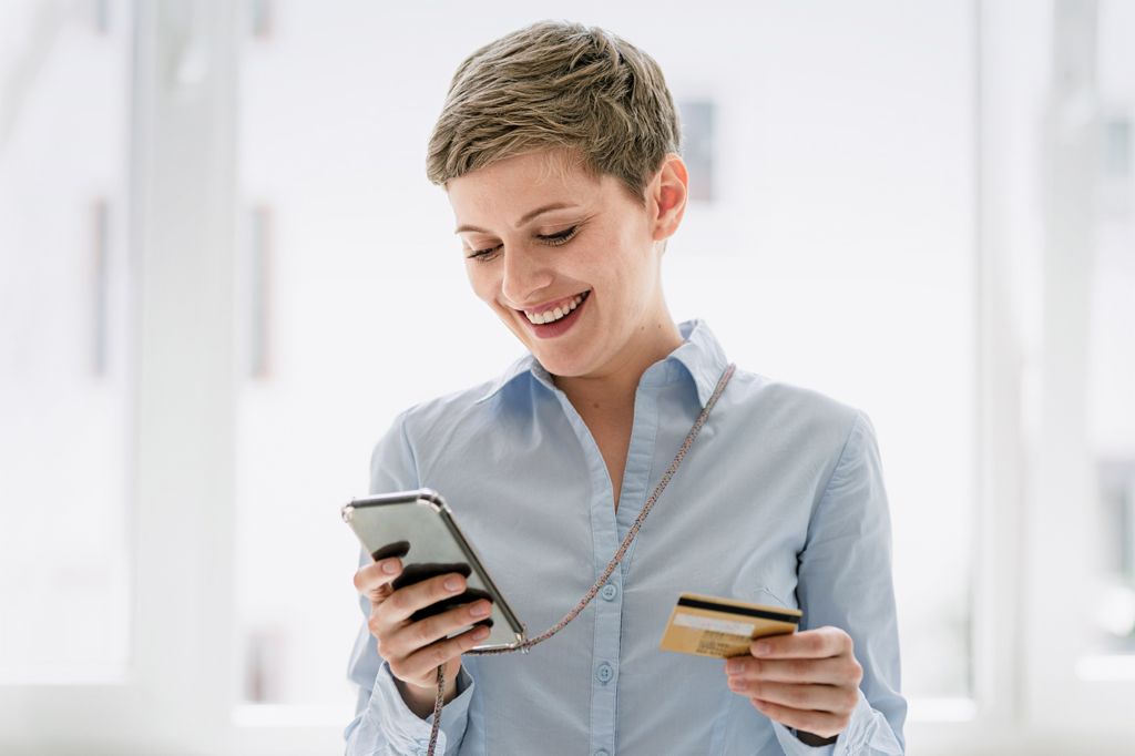 A young woman with short hair wearing a light blue blouse looks at her cell phone and smiles