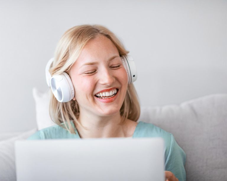 A young woman sits in front of a laptop laughing and wearing headphones