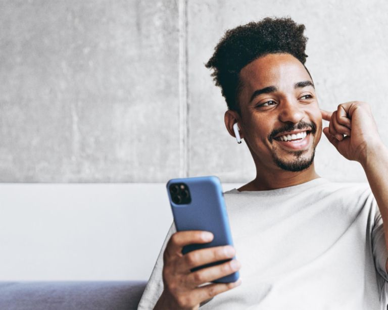 Young fun smiling african american man in beige t-shirt sitting on grey sofa indoors apartment use air pods mobile cel phone listen to music podcast looking aside resting on weekends staying at home