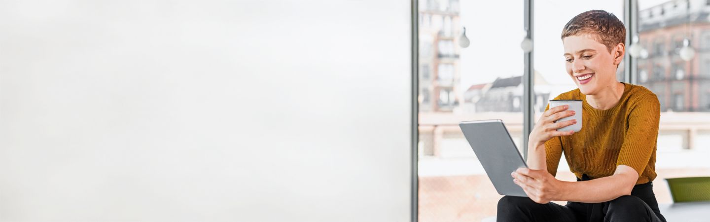Happy woman sitting on table in office with coffee and looking on tablet