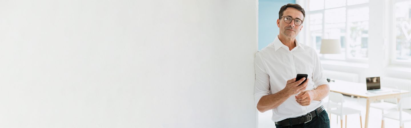 A man in a white shirt leans against a wall, holds his smartphone in his hand and looks seriously into the camera