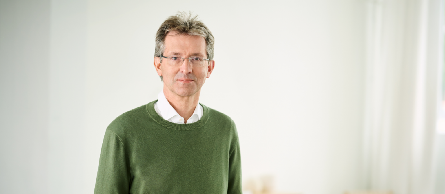 Justus Hecking-Veltman, CFO, mit einem wald-grünen Pullover und weißem Hemd, steht in einem hellen Raum.