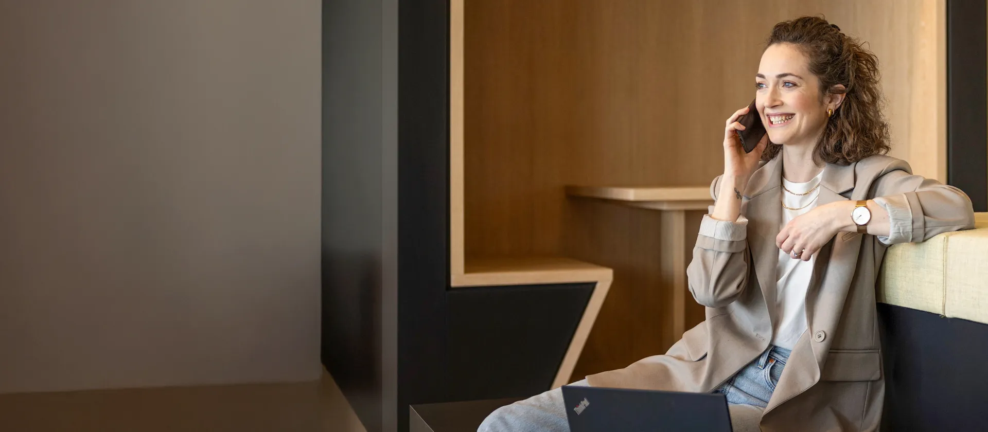 Lächelnde Frau sitzt in einer modernen Büro-Lounge, telefoniert und arbeitet am Laptop.