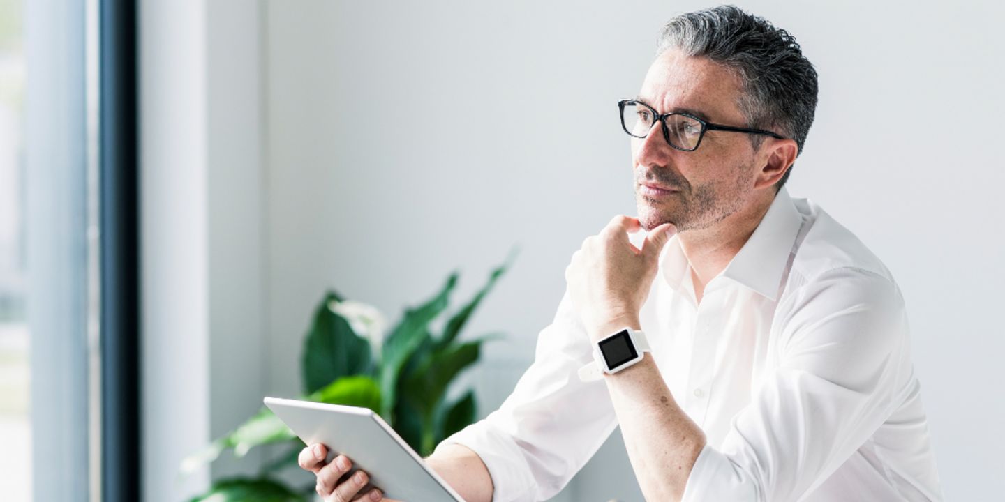 A businessman with glasses sits at his desk in the office with a tablet and thinks.