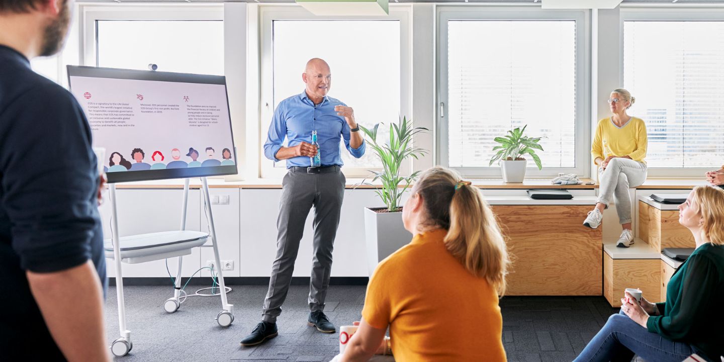 Ein Team in einem modernen Collaborationsbereich eines Büros nimmt an einer Präsentation teil, bei der ein EOS Mitarbeiter vor einem digitalen Whiteboard zu branchenspezifischen Lösungen im Forderungsmanagement spricht.