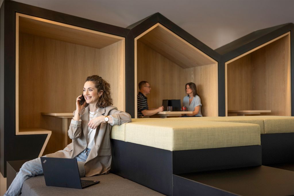 Lächelnde Frau sitzt in einer modernen Büro-Lounge, telefoniert und arbeitet am Laptop. Im Hintergrund arbeiten zwei weitere Personen.