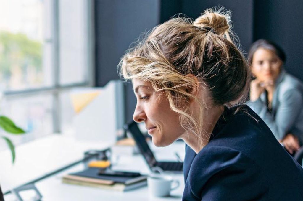 Das Bild zeigt eine Frau, die an einem Schreibtisch in einem modernen Büro sitzt, einen dunklen Blazer trägt und sich auf ihren Laptop konzentriert, während eine Pflanze und einige Büromaterialien auf dem Tisch sichtbar sind und im Hintergrund eine andere Person sitzt.
